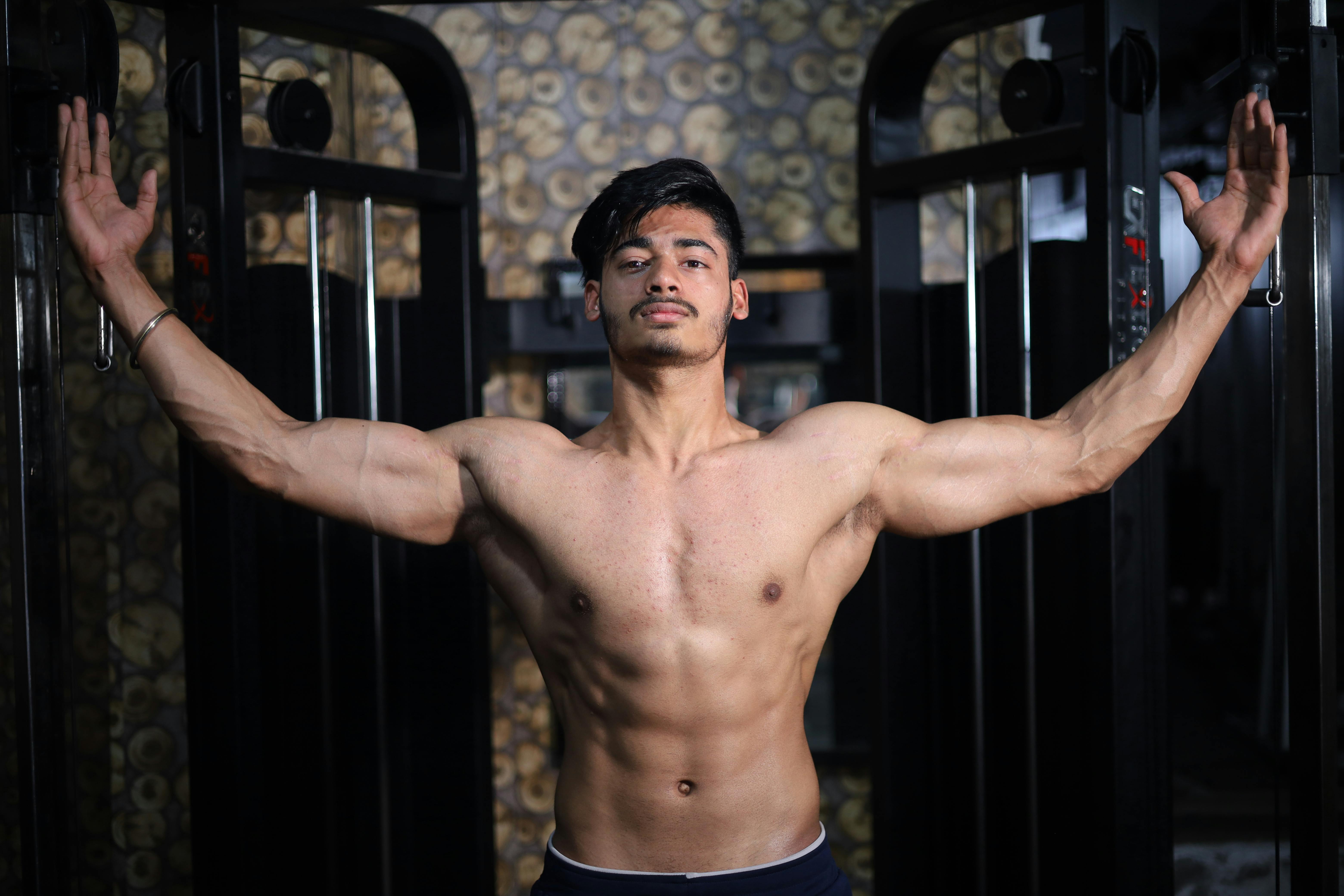 Home A shirtless South Asian man shows off his muscular physique while posing in a gym setting.
